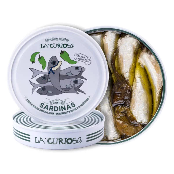 A playful round tin of La Curiosa sardines with green padrón peppers, featuring quirky fish illustrations on the lid. Inside, the neatly packed sardines glisten in olive oil, paired with whole roasted peppers for a bold Galician twist.