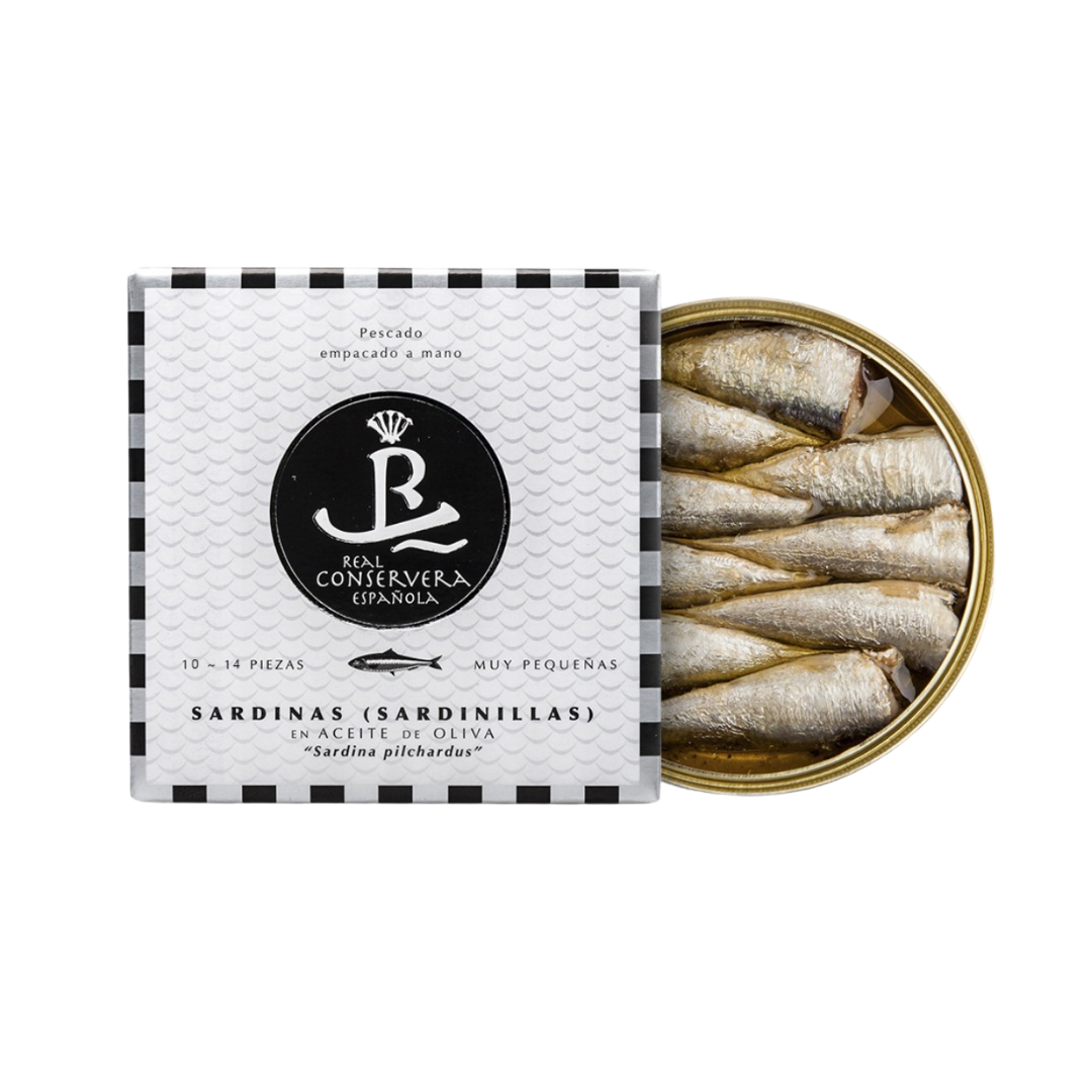 A premium tin of Real Conservera Española Sardinillas reveals delicately packed small sardines in olive oil. The elegant packaging features a black-and-white fish scale design with bold branding, emphasizing artisanal hand-packing and gourmet Spanish seafood tradition.