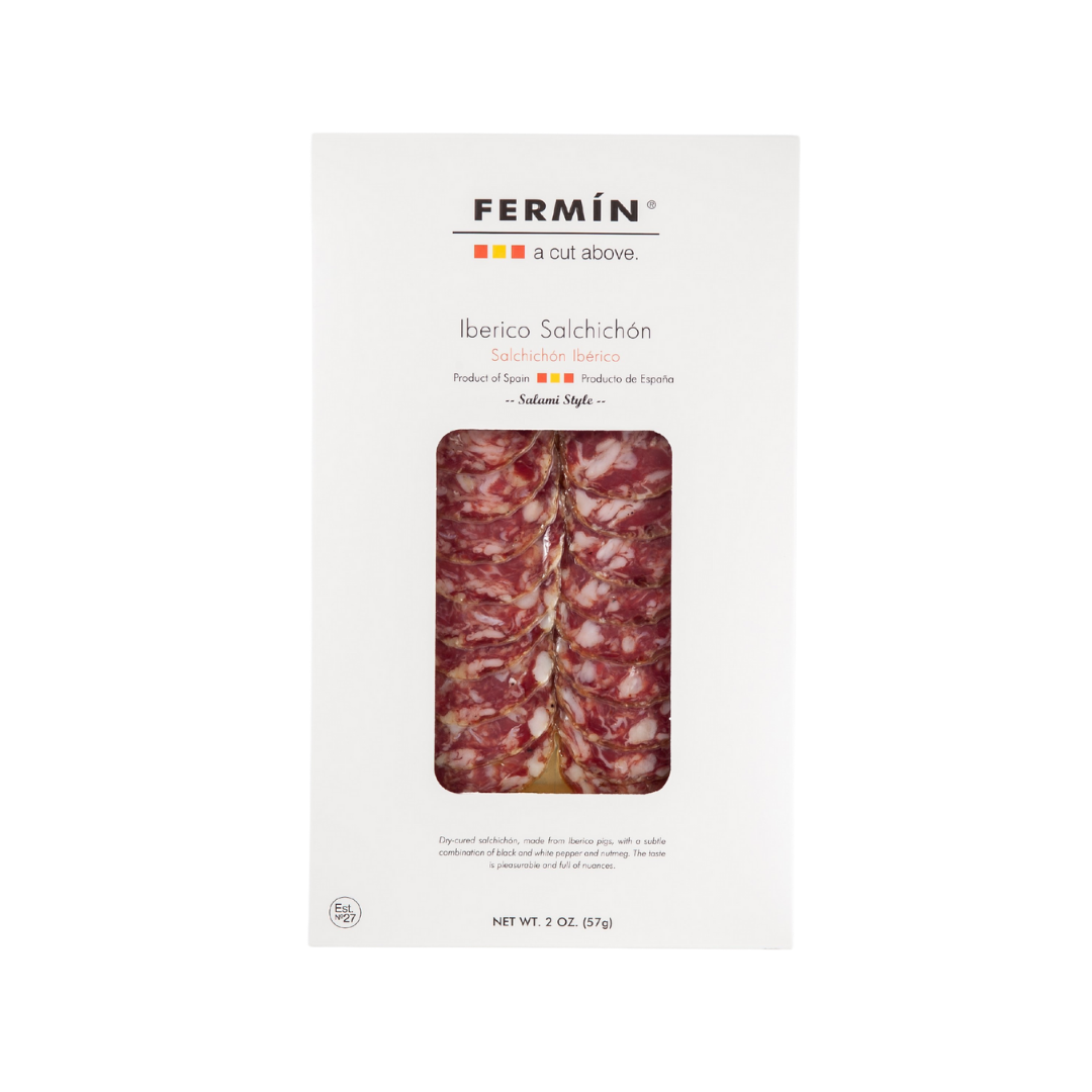 A package of Fermin Iberico Salchichón, a Spanish dry-cured salami made from Iberico pork. The packaging is sleek and white, with a clear window showcasing thinly sliced, marbled salchichón. The label includes the product name in both English and Spanish, along with a brief description noting it contains black pepper and nutmeg. The net weight is 2 oz (57 g).