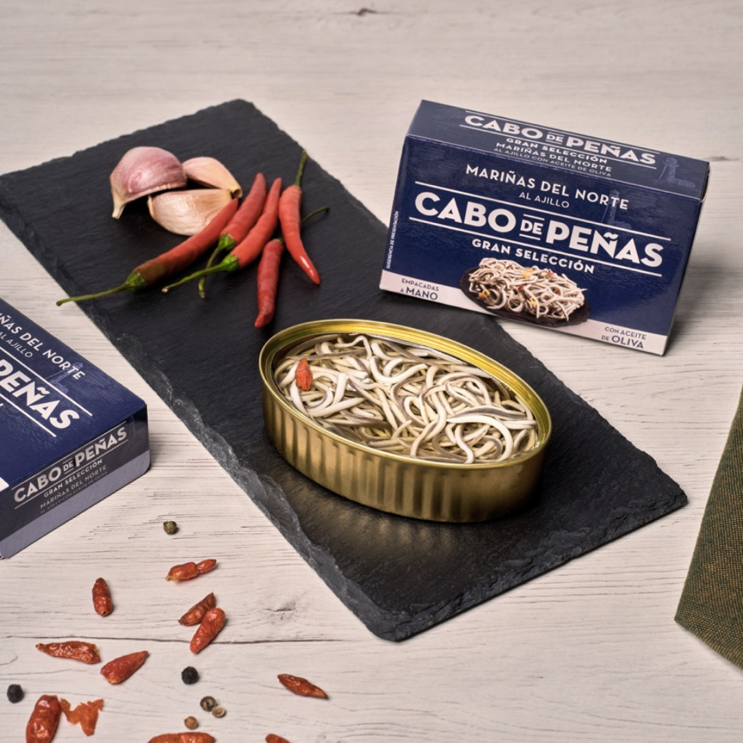 Overhead of a tin of baby eels that is opened, alongside decorative garnishes of garlic and pepper.