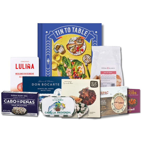 A curated seafood gift set arranged together on a white background, featuring a large blue 'Tin To Table' book or booklet, a Lulína box, a Don Bocarte package, a white bag of fish, a Caballa Provenzal tin, a Cabrito selection box, and other smaller packaged items.